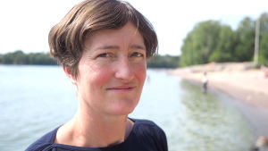 Vivi Fleming-Lehtinen tror inte algläget blir så extremt som i fjol, men varmt väder och svaga vindare kan ännu öka årets algblomning betydligt. Havsforskaren Vivi Fleming-Lindberg står på en strand i Helsingfors och ser mot kameran.