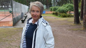 Hangö tennisklubb = Barbro Wikberg, nästan i alla fall. Hon har sysslat med tennis och jobbat för att Hangö tennisklubb ska ha så fina banor och lokaler som möjligt i väldigt många år. En kvinna i vit jacka står nära en tennisbana.