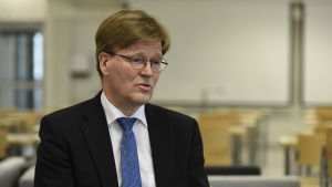 Tapio Kosunen. Tapio Kosunen har mörk kostym, runda glasögon och en blå slips.