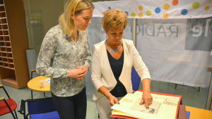 Gia Mellin-Kranck visar reporter Katarina Lind hur Radio Östnylands maskot såg ut förr i tiden. Katarina Lind och Gia Mellin-Kranck tittar på gamla tidningsurklipp.