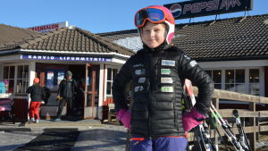 Ellen Fröjdö tycker om att åka i slalombacken. Ellen Fröjdö, en ung flicka med svart jacka, ljusröd hjälm och skidglasögon, står framför en kafeteria på ett skidcenter.