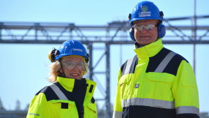Tarja Korvenoja och Arno Avela tycker att plast är ett spännande material att jobba med. Arno Avela och Tarja Korvenoja ler. De har gula jackor på sig och blå bygghjälm på huvudet. Soligt väder.