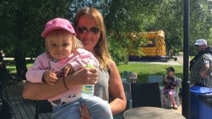 Katja tvivlar inte på att det skulle finnas fästingar också i parkerna i Åbo. Här är hon med dottern Milla. Men ingen av dem har något fästingvaccin. mamman katja håller dottern milla i handen och även en vattenflaska i handen. Milla har en rosa keps och rosa tröja. I bakgrunden syns gröna träd.