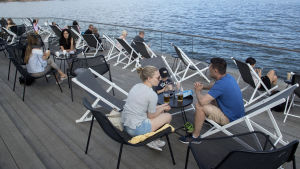 Löyly på Ärtholmen i Helsingfors ägs av Noho Partners, som genast efter att regeringen informerade om nya restriktioner för restaurangerna varslade om samarbetsförhandlingar. Personer sitter på en uteservering i Helsingfors. I bakgrunden syns hav.