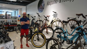 Elykelförsäljaren Mika Sevón tror starkt på elcykelns framtid i Finland. Medelålders man med rakat hår, kortärmad tröja och röda shorts står med armarna i kors i cykelaffär.