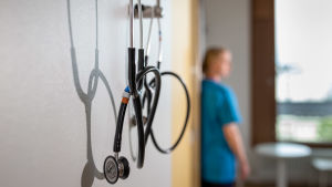 En sjukskötare i ett patientrum. I förgrunden hänger ett stetoskop.