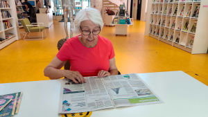 Gun-Britt Eriksson väntar varje vecka på att tidningen ska utkomma. En kvinna i röd skjorta sitter vid ett bord i ett bibliotek och läser en tidning.