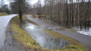 Cykelväg i närheten av Fiskars bruk. En bild på en bilväg och parallellt går en cykelväg. På cykelvägen är det vatten på grund av översvämningar. Runtom vägarna finns träd och brevid cykelbanan vatten.