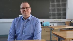 Mikael Kullas är biologi- och geografilärare vid Tegengrens skola i Vörå. mikael kullas sitter i ett klassrum vid en pulpet. i bakgrunden ser man tavlan. kullas bär en blå skjorta och glasögon.