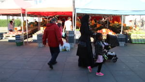 Kvinna med huvudduk promenerar med sina barn på torg med grönskasförsäljning i Tensta.