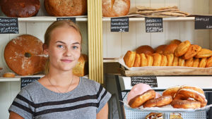 Belinda Zanon hoppas att bilarna skulle sakta ner i och med den nya gårdsgatan. Kvinna står i ett bageri omringad av färska bullar och bröd.