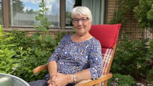 Ann-Mari Svahn sitter i ett av rummen i hennes trädgård. Ann-Mari Svahn sitter i en trädgårdsstol med växter och en gul tegelfasad i bakgrunden.