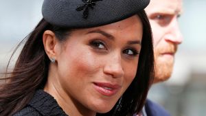 Harry och Meghan träffades genom en gemensam vän som parade ihop dem på en blinddejt. Meghan Markle ler in i kameran med prins Harry i bakgrunden.