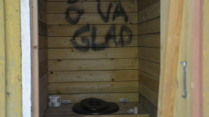 "Skit o var glad" står det i en av utetoaletterna på Ramsholmen. Utedass, problem med vattendistributionen och det faktum att man måste gå en bit till fots är några av utmaningarna för de som vill ordna fester på Ramsholmen idag. Ett utedass med svart toalettsits och lock av styrox. rätt okej, lite smutsigt. På väggen bakom toalettringen står det med spraymålfärg "Skit o va glad".