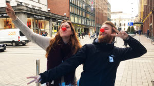 Milli Hellström och Christoffer Kaski iförda röda näsor framför en gata med människor som går.