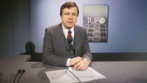 Uutisankkuri Arvi Lind käsittelemässä tupo-aihetta. Yleisradion TV-uutiset. Toimittaja Arvi Lind lukemassa uutisia tv-studiossa, taustalla TUPO-aiheinen dia (tv-grafiikka, taustakuva). Uutisankkuri työssä. Uutistoimittaja juontaa tv-lähetystä. Uutisstudio. Yleisradion ohjelmat.
