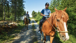 Kyösti Linna rider på hästen Sockan och bakom kommer André Norrgård på Laasi. Två män rider på varsin häst längs en skogsväg.