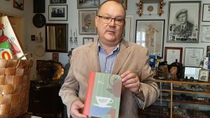Mikael Andersson och boken om kaffe. man i ljus kostym och blå skjorta och glasögon står och håller i bok om kaffei rum fullt av gamla föremål