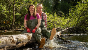 Minna Pyykkö ja Pirkka-Pekka Petelius istuvat puunrungolla joella.