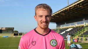 Lewis Thomas är Forest Green Rovers målvakt. En glad fotbollsspelare i en ljusröd tröja. Han står på en gräsplan vid en tom läktare.