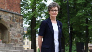 För Marja Routi är Åbo domkyrka förutom en arbetsplats också en speciell och helig plats. En kvinna med brungrått hår och glasögon är klädd i en mörkblå kostymväst och en vit t-skjorta, och står på Åbo domkyrkas trappor med träd i bakgrunden.
