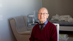 Eero Halonen on asunut pienessä evakkoyksiössään jo yli vuoden. Eero Halonen istuu ja katsoo kameraan. Taustalla näkyy nojatuoli ja sijaamaton vuode.