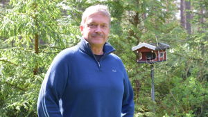 Torbjörn Ekholm (SFP) är stadsstyrelseordförande i Hangö. En man i blå tröja poserar i en grön, skogsliknande trädgård. Bakom finns ett fågelbräde som är som ett rött hus.