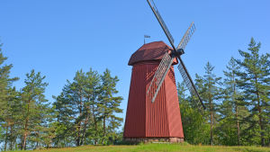 Den vackra kvarnen ska få en nya lekpark som granne. En röd kvarn står på en gräsmatta omgiven av tallar.