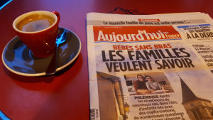 I går toppade stora dagstidningen Aujourd’hui med titeln ‘Familjerna vill veta’ och har gjort stora reportage om barnen som fötts utan armar eller händer. En bild på en tidning, bredvid finns en röd kaffekopp.