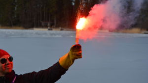 Frihetsgudinnan Mikael Furu. En man med röd mössa och solglasögon håller i ett nödbloss som brinner med en rödorange färg.