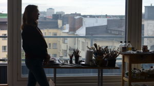 Målaren Magdalena Åberg vid stort fönster i motljus. i fönstret syns många hustak och himmel, på bordet framför fönstret penslar och färger