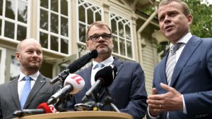 Europa- och kulturminister Sampo Terho (Blå), statsminister Juha Sipilä (C) och finansminister Petteri Orpo (Saml) höll en kort presskonferens inför budgetmanglingen. Regeringstrion står uppställd med allvarliga miner klädda i kostym med mikrofoner framför sig. I bakgrunden Villa Bjälbo.