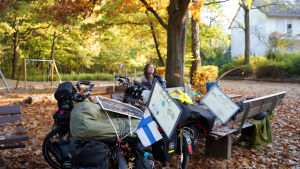 Den tunna plattan ovanpå väskan är en solpanel. En kvinna sitter på en bänk i en park. Det är höst och det ligger gula löv överallt. I förgrunden syns en cykel som har en massa väskor på sig samt Finlands flagga.