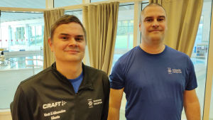 Idrottsinstruktör Patrik Nyberg (till vänster) och hallmästare Benny Forsström är båda glada över att Ekenäs simhall har öppet igen. Portättbild på två män, Patrik Nyberg och Benny Forsström, klädda i sportjacka respektive t-skjorta. Bakom stora fönster (i bakgrunden) ser man Ekenäs simhall med sina bassänger.