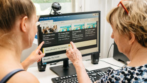 Eläkeikäinen nainen ja häntä digitaidoissa auttava nuorempi sukulainen tietokoneen äärellä, kädet osoittavat koneen näyttöä.