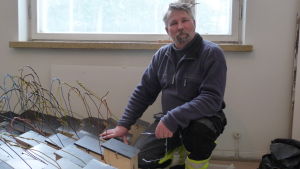 Tom Lindgren, arbetshandledare, med en del av vinterns produktion. Tom Lindgren knäböjer framför omålade fågelholkar. Inomhus, ett fönster bakom. .