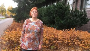 Liisa Harjula utanför Valonia Medelålders rödhårig kvinna i brunrödgrå blus ler framför buskar och tegelbyggnad