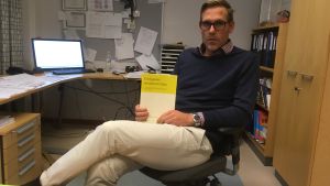 Peter Boström och nya handboken för kvalitetsvård av urologiska sjukdomar lång man i mörkblå tröja och rutig skjorta och ljusa byxor och glasögon sitter i sitt arbetsrum på en kontorsstol och håller en bok i sin hand