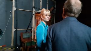 Janina Andersson och Erkki Liikanen blond kvinna med glasögon och uppsatt år och ljusblå jacka står och talar med man i kostym som man bara ser bakifrån. Rummet är mörkt