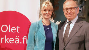 Janina Andersson och Erkki Liikanen Ung kvinna med glasögon och uppsatt blont hår. Hon har på sig en ljusblå kavaj. Bredvid henne en man i övre medelåldern med kostym, vit skjorta, kravatt, glasögon
