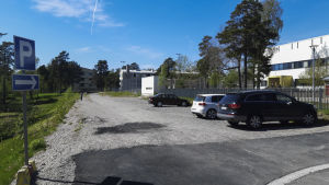 Den nuvarande planen är att paviljongen ska stå på den här grusplanen. Den ligger mellan den finska lågstadieskolan och några ambassader. En grusplan. På höger sida syns ett hus med en gårdsplan där barn leder. Några bilar står parkerade på grusplanen.