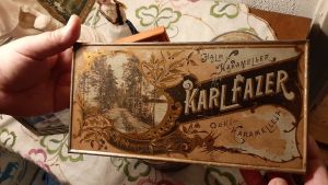 Halmkarameller. två händer håller i en mycket gammal ask med halmkarameller från Karl Fazer. färgerna skriftar mellan ljust och mörkbrunt