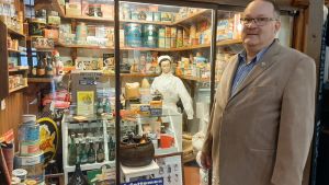 Mikael Andersson framför sin museibod. man i glasögon och ljus kavaj står framför museibutik med gamla livsmedelsförpackningar och skyltdocka