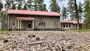 Askersgården måste rivas för att ge utrymme för det nya skolcentrumet. I huset har det funnits rådgivning och bibliotek. Huset är ett av dem som svenskarna donerade efter kriget, därav namnet Askersgården. En bild på en grå byggnad i rött tak. I förgrunden syns barr och kottar.