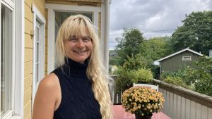 Janina Andersson. En kvinna med långt, blont lockigt hår står vänd mot kameran och ler. Hon står på verandan av ett gult trähus, och bakom henne syns buskar och träd.
