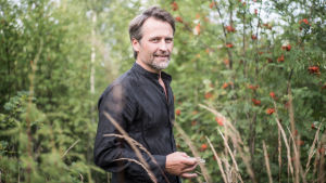 Jan Söderblom, som bor i Fiskars med sin familj, säger att tystnaden och naturen är hans inspirationskällor. En man i svart läderjacka med grått skägg står i naturen med bland annat röda rönnbär i bakgrunden. Han står snett så att han vänder huvudet mot kameran med ett litet leende på läpparna.