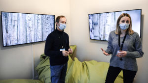 Ledande psykolog Tero Timonen och klinisk specialsjukskötare Anne Priha visar ett av ätstörningsenhetens nya konstrum, med Johanna Ketolas videoverk. En man och en kvinna i munskydd står inne i ett rum och berättar något mot kameran. I bakgrunden syns stora gröna kuddar och två skärmar på väggen som visar naturbilder.