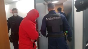 Den yngre kvinnan anländer till häktingsförhandlingen. En person i röd rock med huvan på ledsagas till en rättegångssal av två väktare.