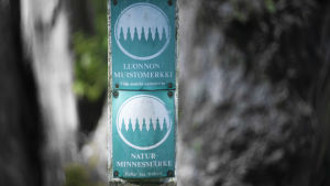 En skylt som det står "Naturminnesmärke" på med en illustration av en skog. I bakgrunden syns ett träd.