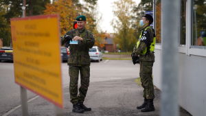 Mika Immonen berättar att han själv alltid har ett munskydd i fickan han kan sätta på sig vid behov. Två personer i uniform står utomhus vid ett vaktbås och tittar på varandra. Den ena har munskydd och den andra håller ett munskydd i sin hand.